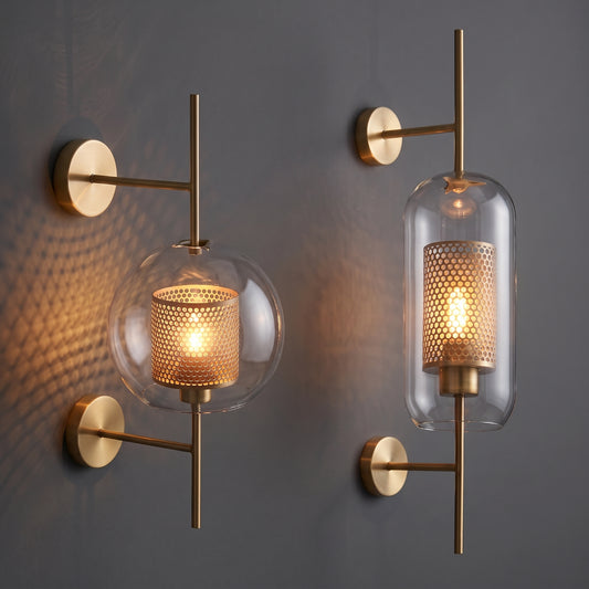Two modern wall sconces with brushed brass hardware and clear glass shades, one spherical and one cylindrical, mounted on a dark gray wall with warm light reflections.