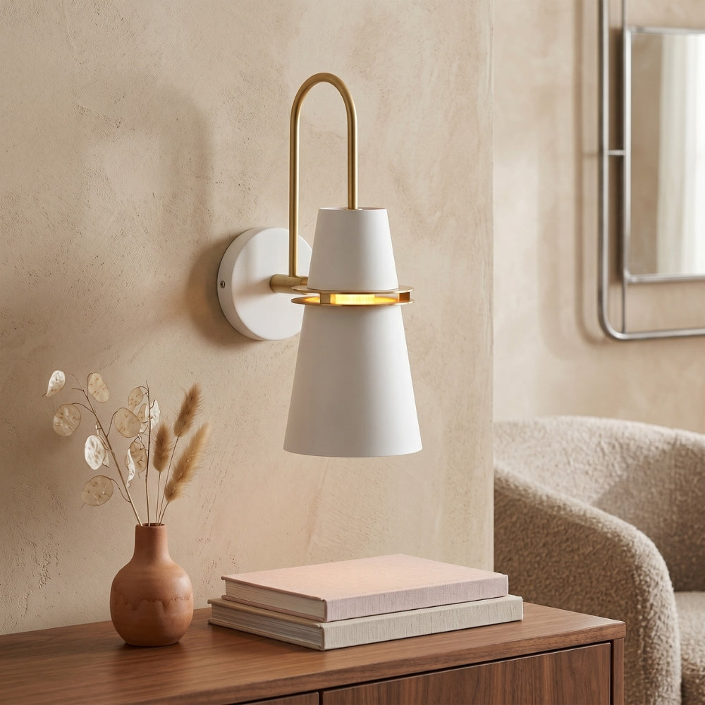 Modern wall sconce with white shade and gold accents on a beige wall, with books and a vase on a wooden surface below.
