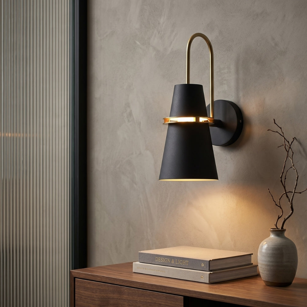 Modern wall sconce with black shade and gold accents on a textured wall above a wooden surface with books and a vase.