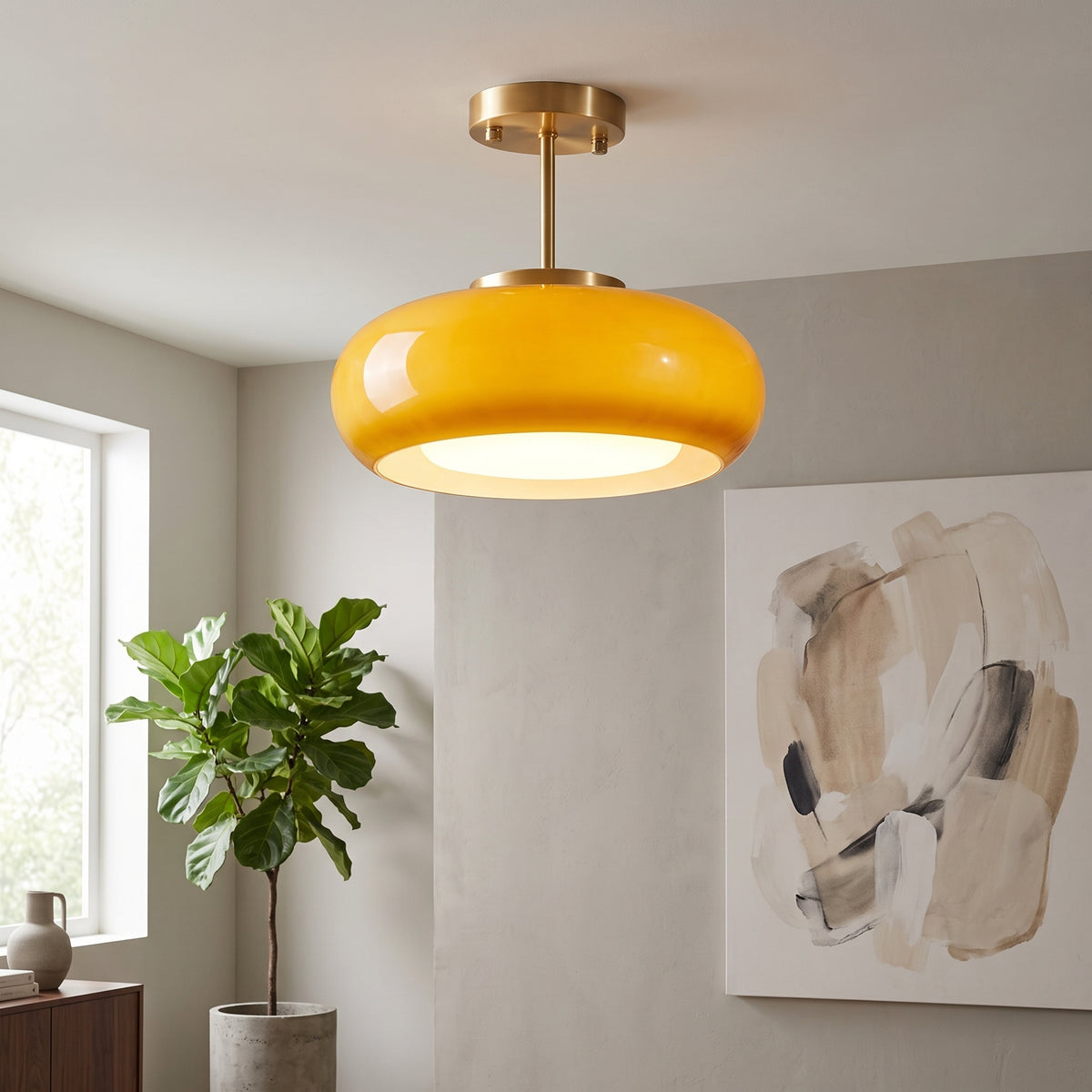 A modern semi-flush mount ceiling light with a glossy yellow-orange dome shade and a brushed brass metal stem, shown hanging in a styled room with a potted plant and abstract wall art.