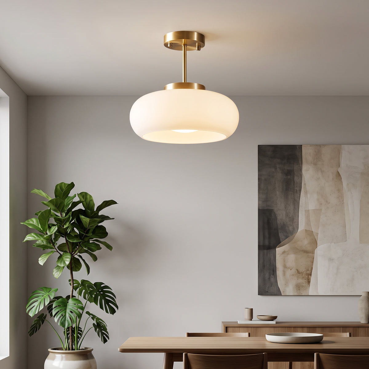 A modern semi-flush mount ceiling light with a warm brass finish and a large, rounded white frosted glass shade, shown hanging in a minimalist dining room with a potted plant and wooden table.