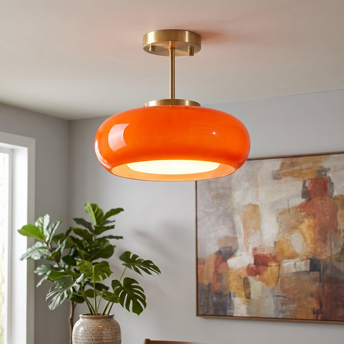 An orange glass pendant light with a gold-toned ceiling canopy and stem, hanging in a modern room with a potted plant and abstract wall art.