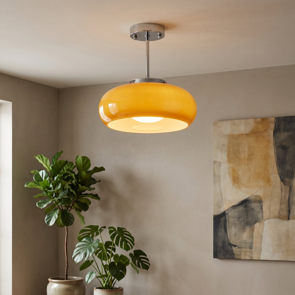 A modern pendant light with a glossy amber-yellow dome shade and a brushed silver metal canopy and stem, hanging in a styled room corner with potted plants and a wall art piece.