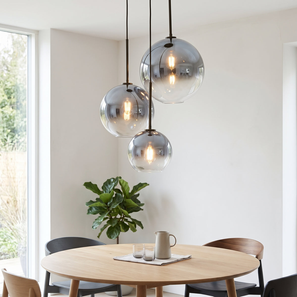 Three modern glass pendant lights with dark metal hardware hang over a round wooden dining table in a bright, contemporary room.
