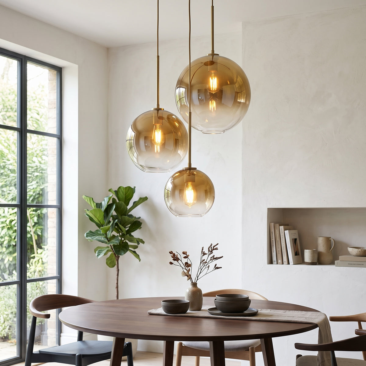 Three amber-tinted glass pendant lights hang over a round wooden dining table in a bright, modern dining room with white walls, a built-in shelf, and a potted plant.