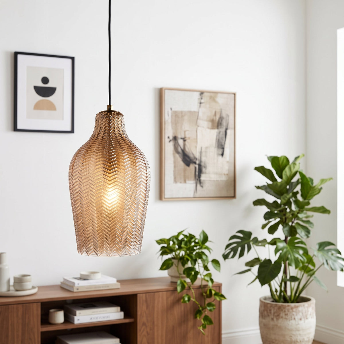 A woven pendant light with a warm glowing bulb inside, hanging in a bright, modern room with framed wall art, potted plants, and a wooden console table.
