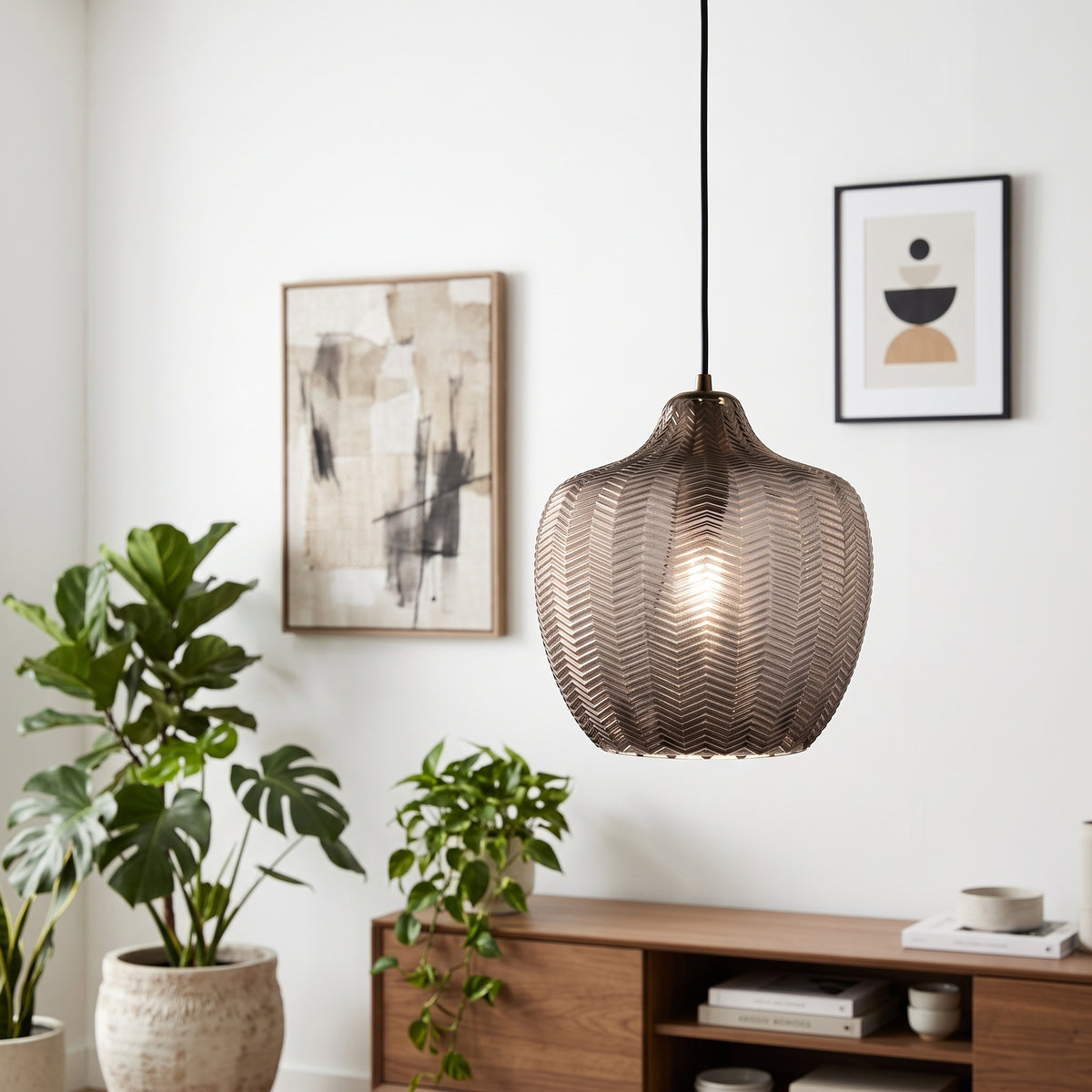 A modern pendant light with a textured, woven metallic shade in a bronze finish, hanging in a styled living room setting with plants and framed wall art.