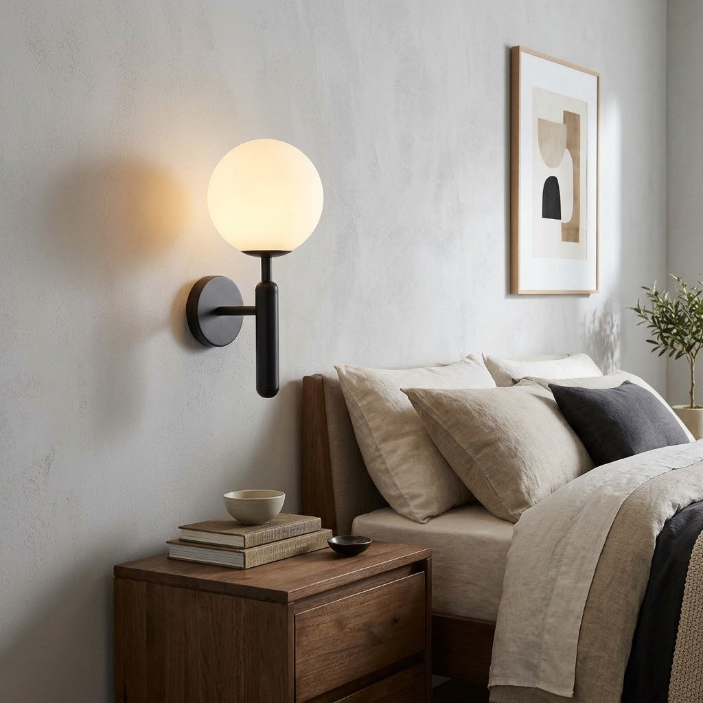 A modern wall-mounted spherical globe lamp with a black metal mount and warm glowing light, shown installed on a bedroom wall above a wooden nightstand and bed.