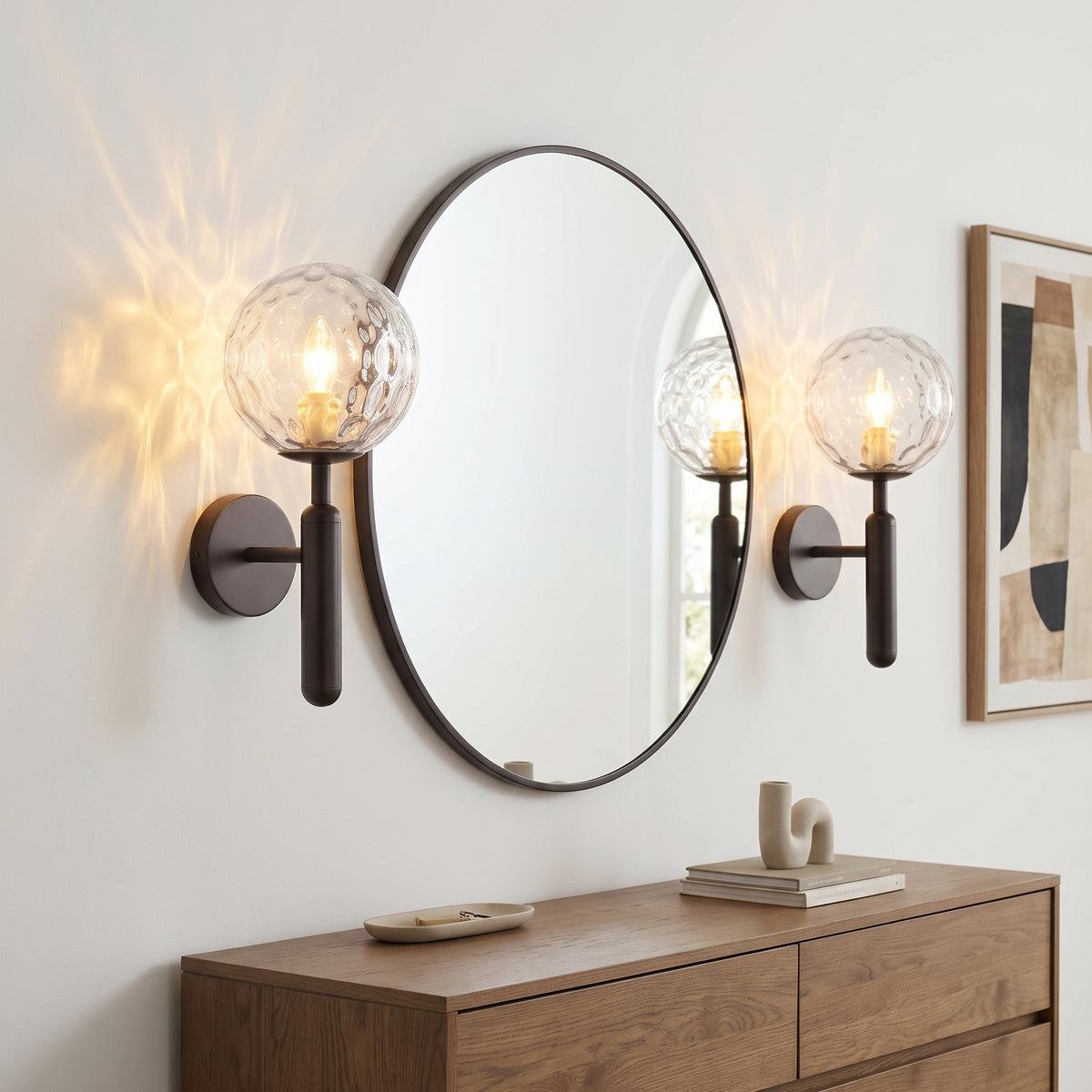 A pair of modern wall sconces with clear textured glass globes and dark metal mounts, installed on either side of a large oval wall mirror above a wooden console table.
