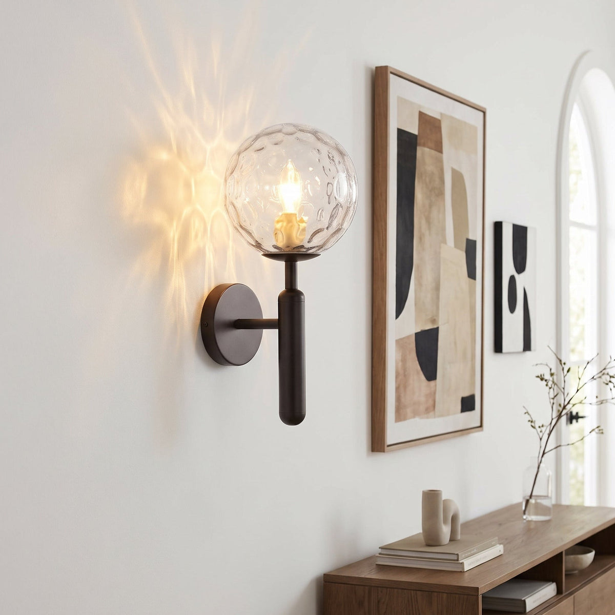 A modern wall sconce with a clear textured glass globe and a dark metal stem, mounted on a white wall in a styled interior next to framed abstract art and a wooden console table.