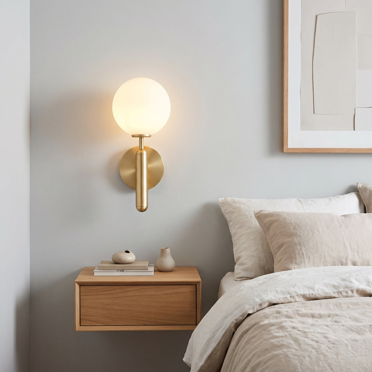 A modern wall-mounted light fixture with a frosted white spherical globe and brushed brass cylindrical stem, installed on a bedroom wall above a floating wooden nightstand next to a bed.