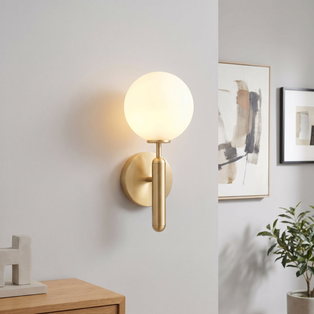 A modern wall-mounted light fixture with a spherical frosted glass globe and a brushed brass cylindrical stem, shown lit on a white wall in a styled interior setting.