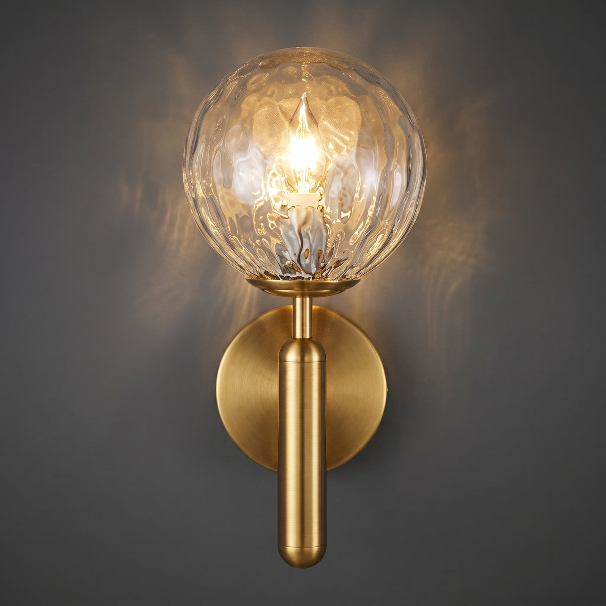 A modern wall sconce light fixture with a textured clear glass globe shade and a brushed gold metal stem and circular wall mount, shown illuminated against a dark gray background.