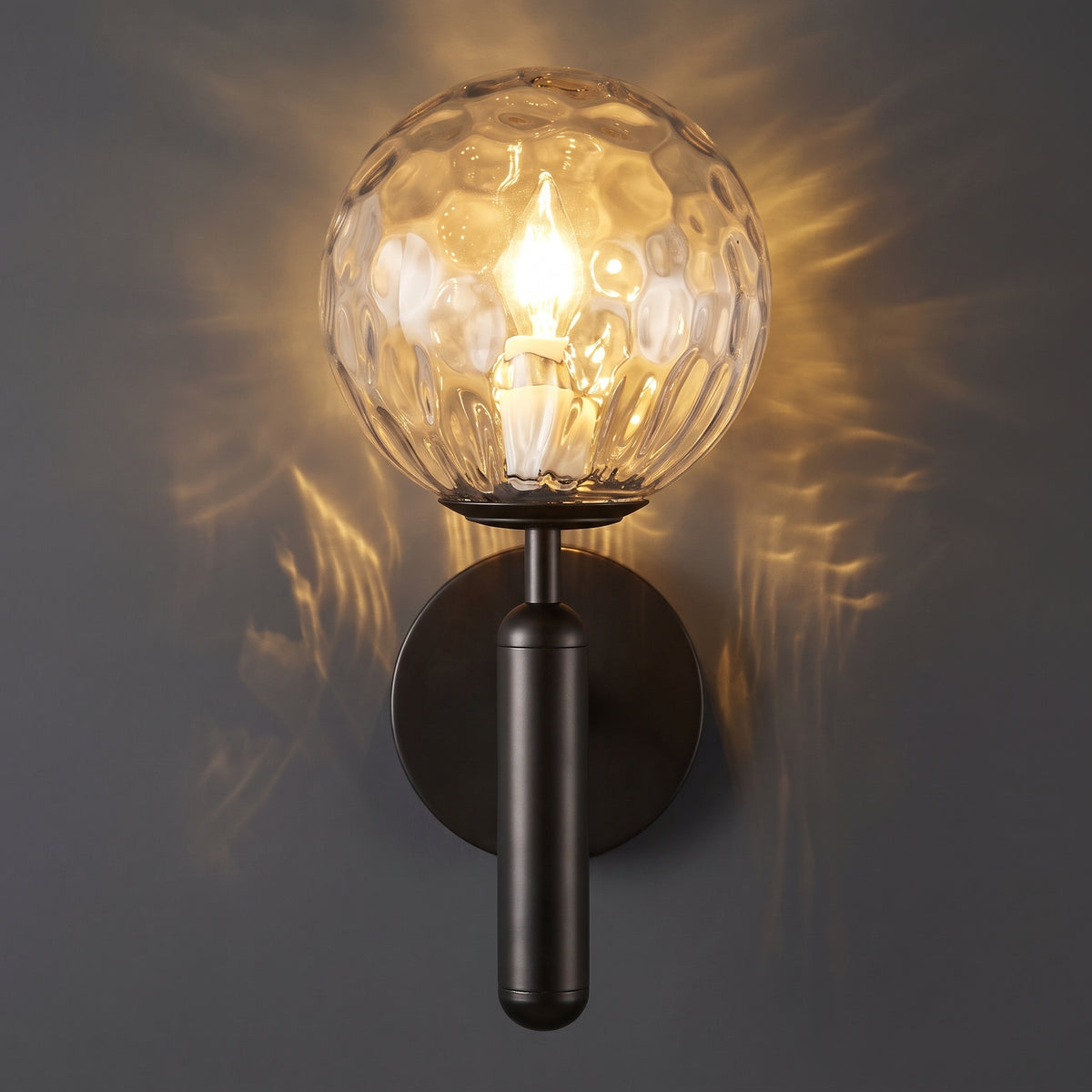 A modern wall sconce with a clear textured glass globe shade and a dark metal cylindrical stem and round wall mount, shown lit against a dark gray wall with warm light rays.