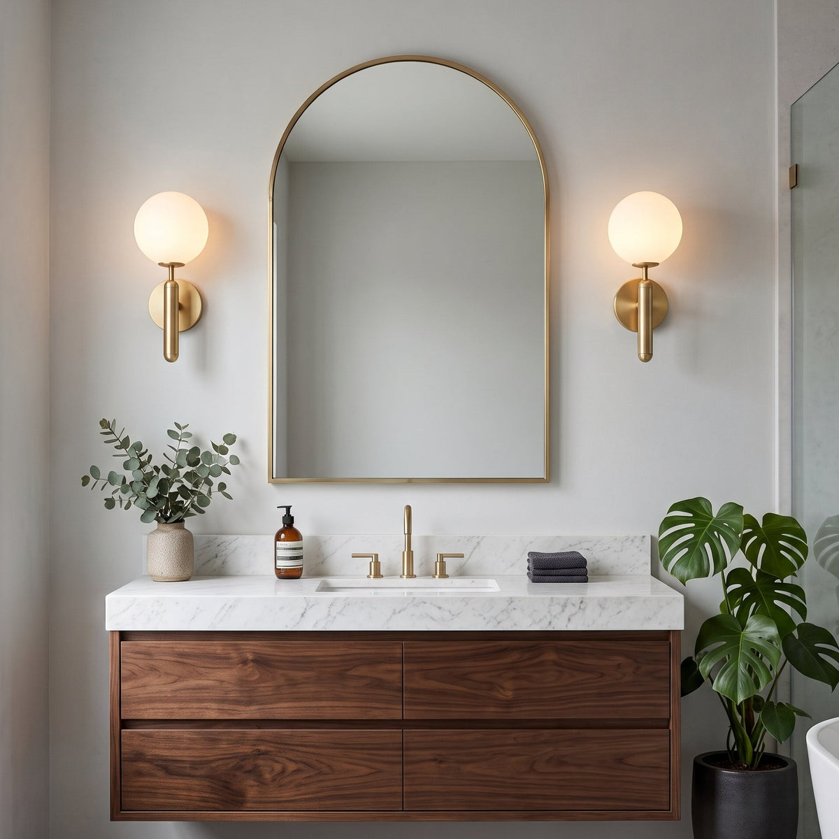 A modern floating bathroom vanity with a white marble countertop, dark wood cabinet, and an arched gold-framed mirror. Two gold wall sconces with glowing globe bulbs are mounted on the light gray wall on either side of the mirror.
