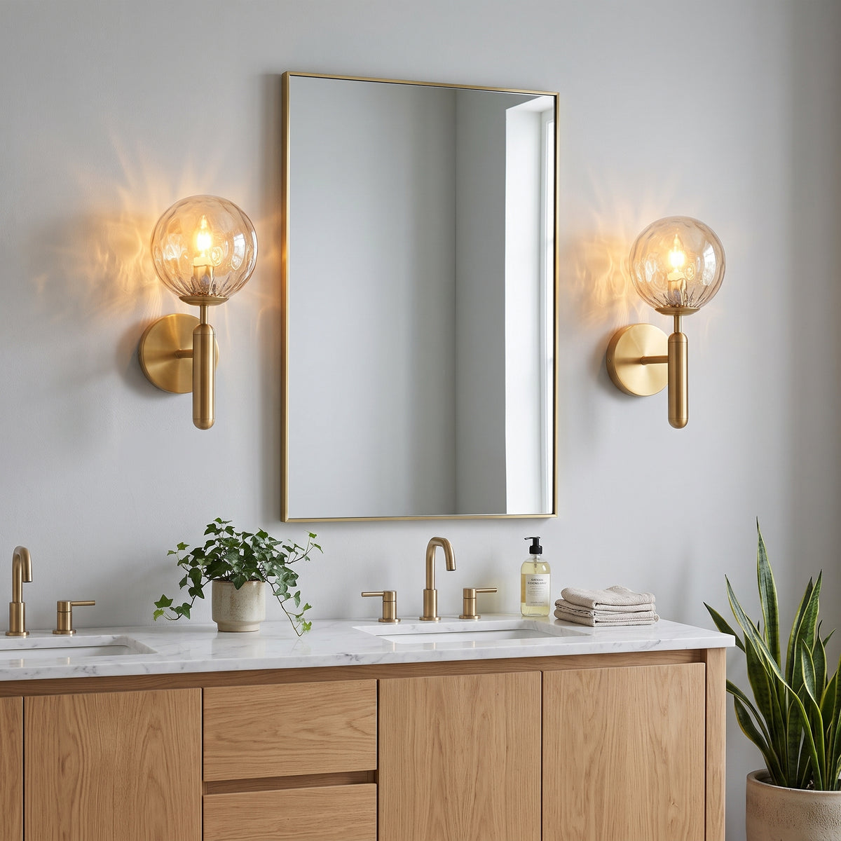 A modern bathroom scene with a rectangular mirror, two matching gold wall sconces with clear globe bulbs, a light wood vanity, white marble countertop, and potted plants.