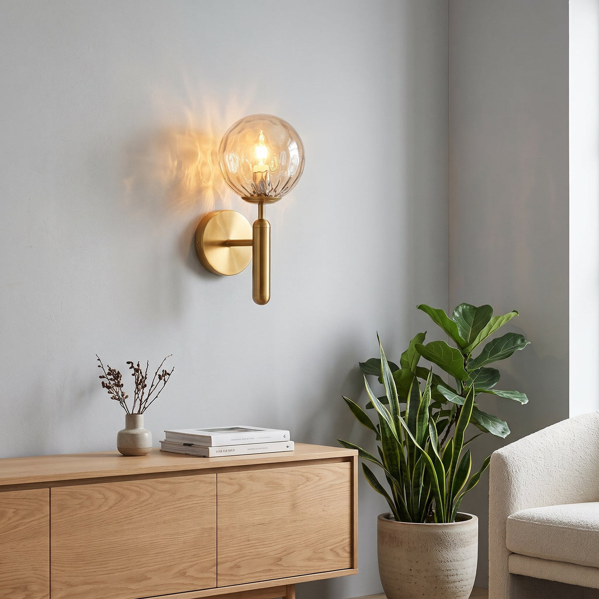 A modern wall sconce with a warm brass-finished metal mount and a clear glass globe shade, glowing with a warm light. It is mounted on a light gray wall above a light wood console table with books and a small vase, next to a potted green plant and a light-colored armchair.