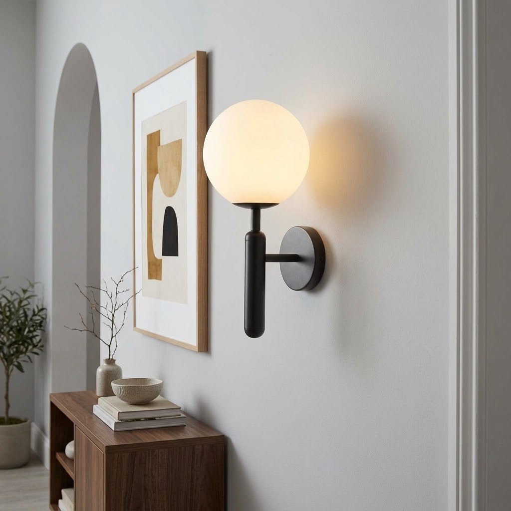 A modern wall-mounted light fixture with a glowing spherical frosted globe and a matte black metal arm and circular wall mount, shown installed on a white wall in a styled interior with framed art, a wooden console, and a plant.