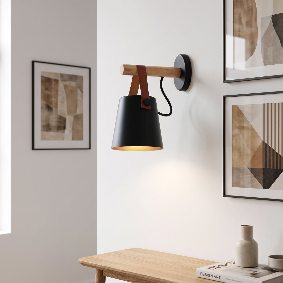 A modern wall-mounted swing-arm lamp with a matte black cone shade, light wood arm, and brown leather strap details, shown lit in a styled interior setting with framed wall art and a wooden table.