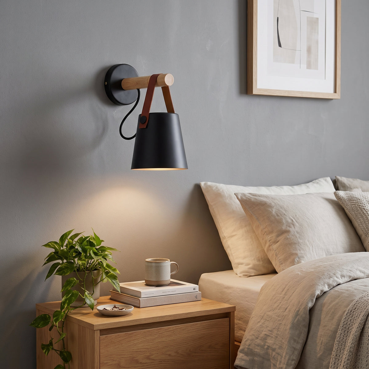 A modern wall-mounted lamp with a matte black cone shade, light wood arm, and brown leather straps, shown lit on a gray bedroom wall above a wooden nightstand.