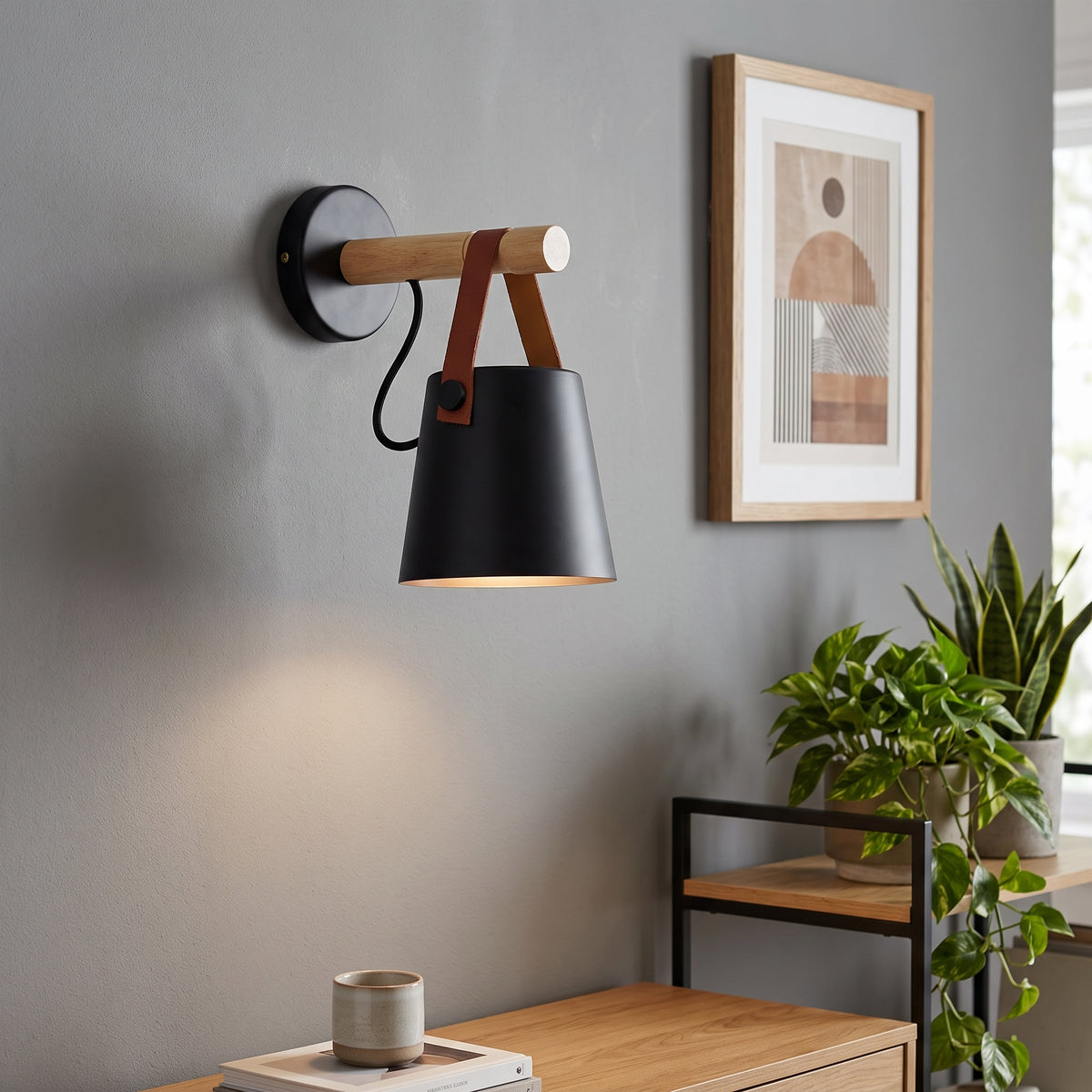 A modern wall-mounted sconce with a matte black cone-shaped shade, a light wood arm, and brown leather straps, shown lit on a gray wall in a styled interior with framed art and a plant.