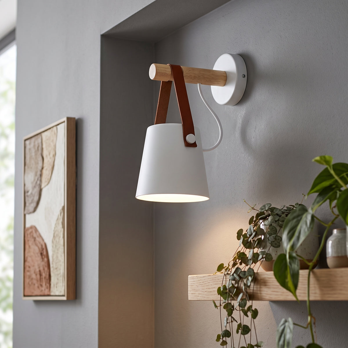 A modern wall-mounted sconce with a white cone-shaped shade, a light wood arm, and brown leather straps, shown lit on a gray wall in a styled interior with framed art and a plant.