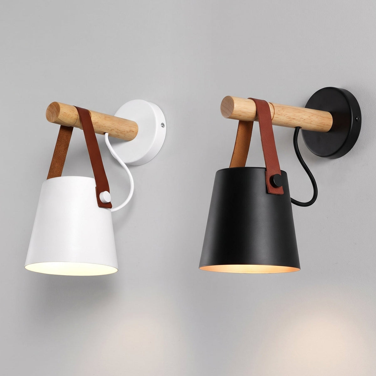 Two modern wall sconces shown side by side against a light gray wall. The left sconce is matte black with a natural wood rod and brown leather strap, and the right sconce is white with the same wood rod and brown leather strap.