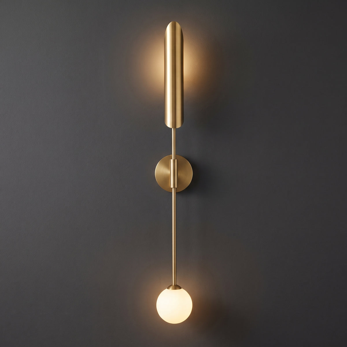 A modern wall-mounted sconce light fixture in brushed brass with a vertical cylindrical shade, a central stem, a circular wall mount, and a glowing spherical bulb at the bottom, shown against a dark gray wall.