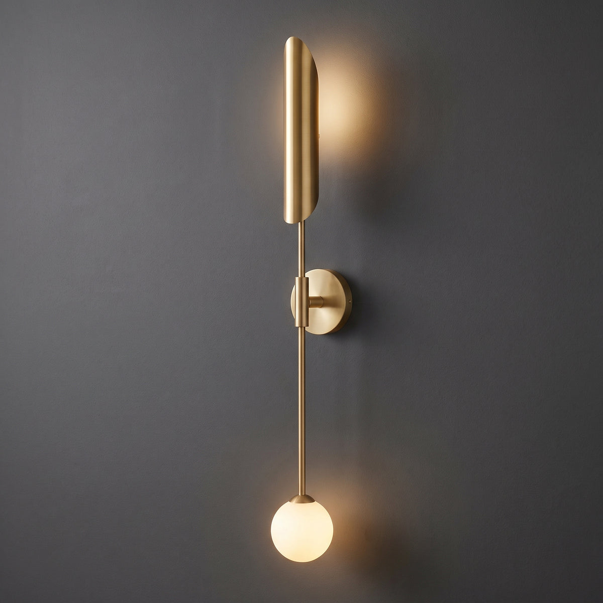 A modern wall sconce with a brushed brass/gold finish, featuring a vertical cylindrical shade, a thin central rod, a circular wall mount, and a glowing frosted spherical bulb, shown against a dark gray wall.