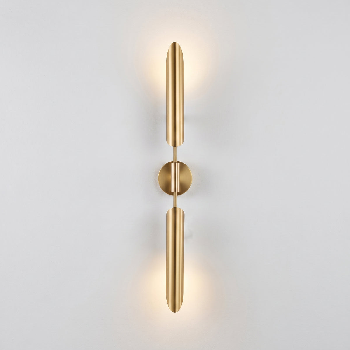 A modern wall-mounted light fixture with two elongated cylindrical arms connected by a round center plate, shown lit against a plain light gray wall.