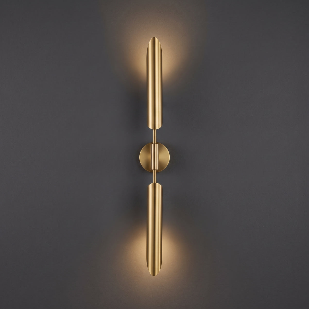 A modern wall-mounted light fixture with two elongated cylindrical arms in a brushed gold finish, mounted on a dark gray wall and casting a warm glow upward and downward.