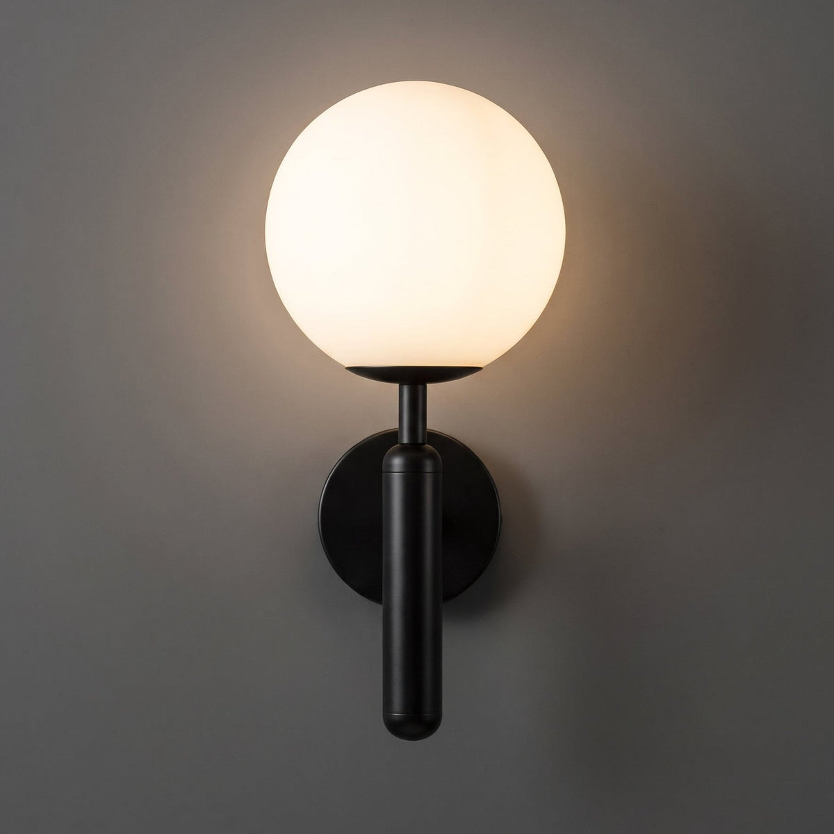 A modern wall-mounted sconce with a glowing frosted white spherical globe and a matte black metal stem and wall mount, shown against a dark gray wall.
