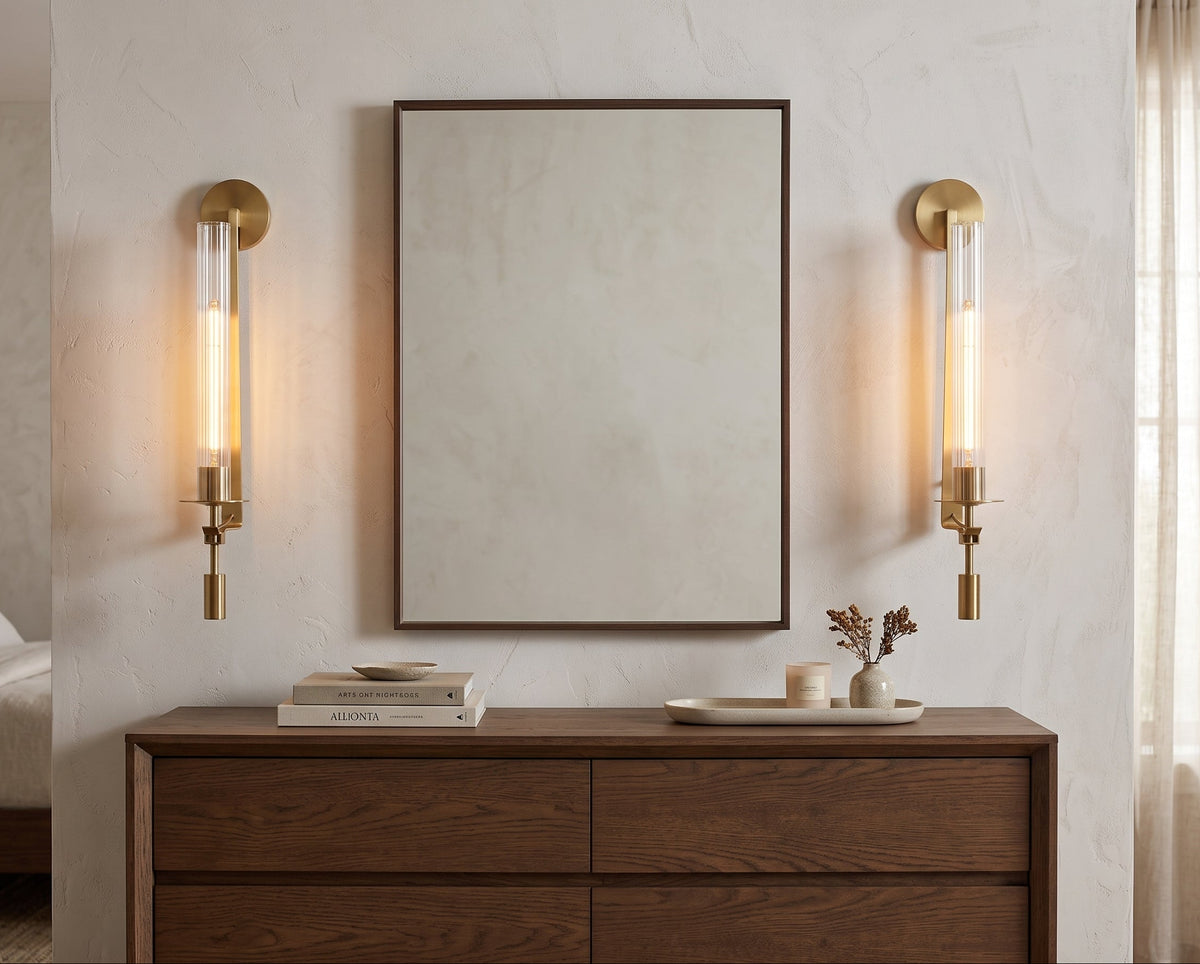 A modern bedroom scene featuring a large square wall mirror with a thin dark frame, flanked by two matching brass wall sconces with clear glass tubes. Below the mirror is a dark wood dresser with books and small decorative items.