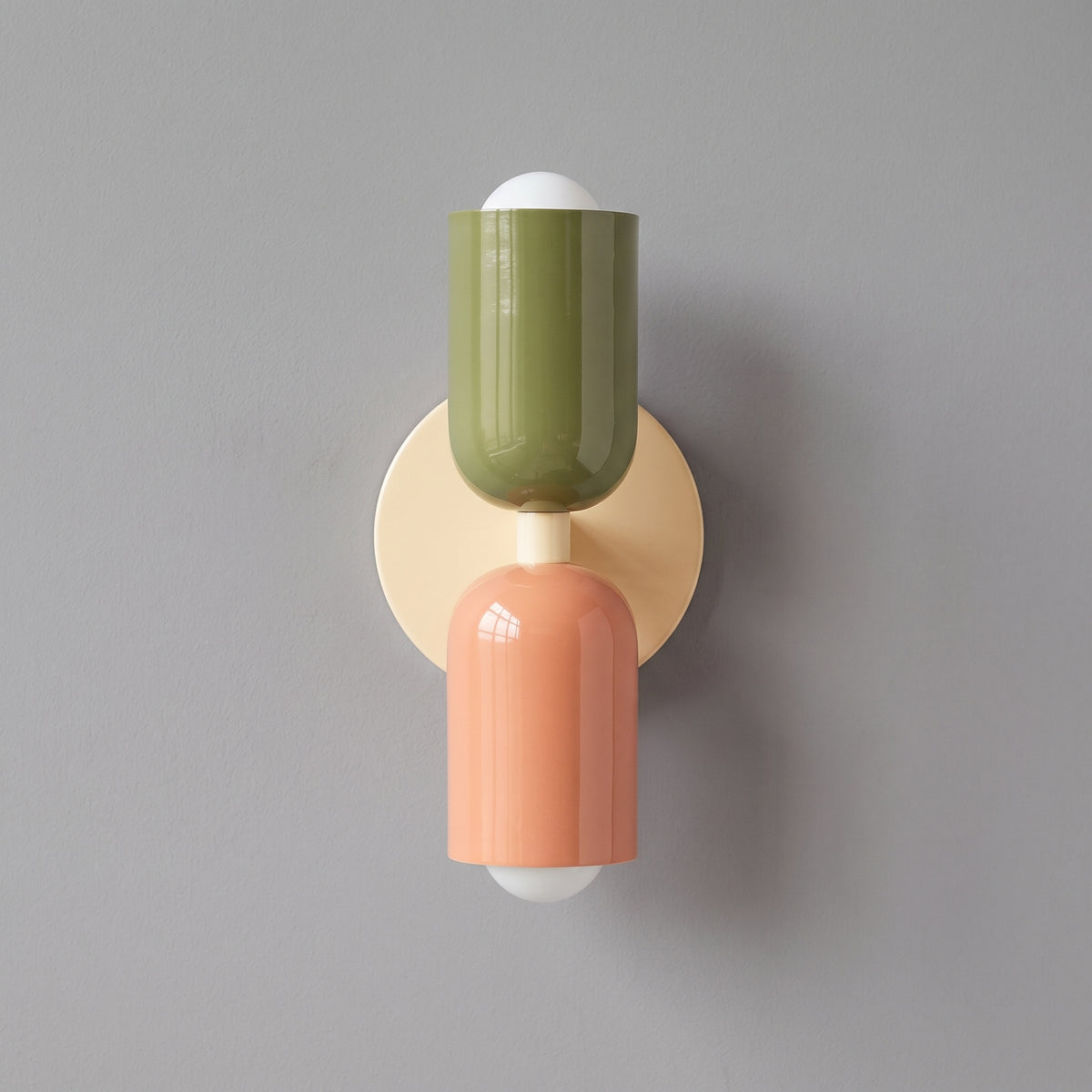 A modern wall-mounted light fixture with a round beige backplate, featuring two glossy cylindrical shades in olive green and peach pink, with white globe bulbs at the top and bottom, set against a solid gray wall.