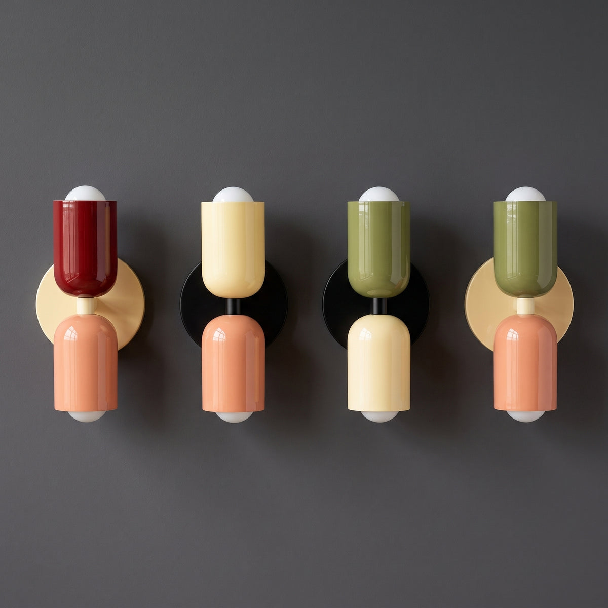 Four modern wall-mounted light fixtures arranged in a row against a dark gray wall, each with a round backplate and glossy cylindrical shades in red, cream, peach, olive green, and peach.