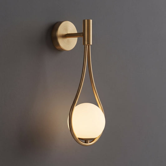A modern wall-mounted light fixture with a brushed brass/gold finish and a glowing frosted glass globe, shown against a dark gray wall.