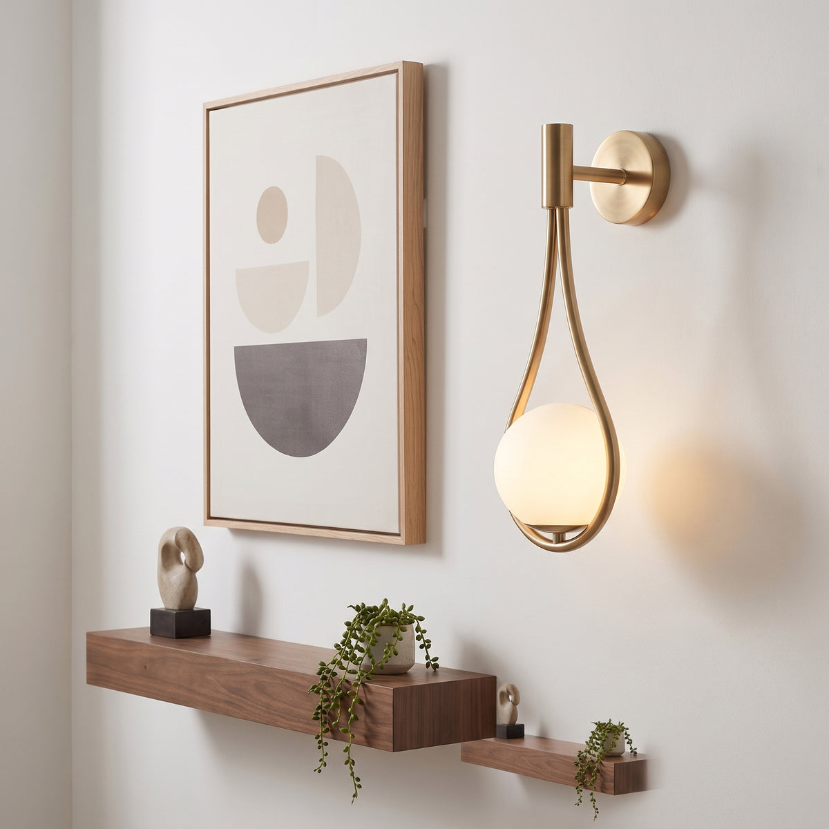 A modern wall-mounted sconce with a gold-toned metal frame and a glowing frosted glass shade, shown on a white wall beside framed abstract art and floating wooden shelves.