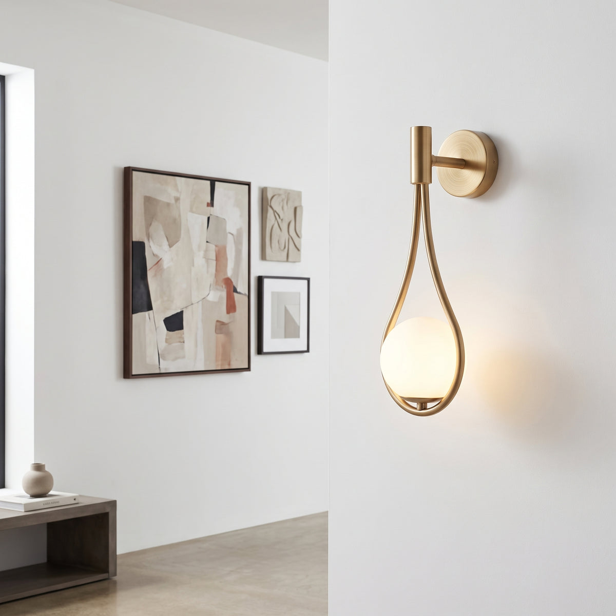 A modern wall-mounted light fixture with a brushed gold metal frame and a glowing frosted glass teardrop shade, installed on a white wall in a minimalist interior setting.