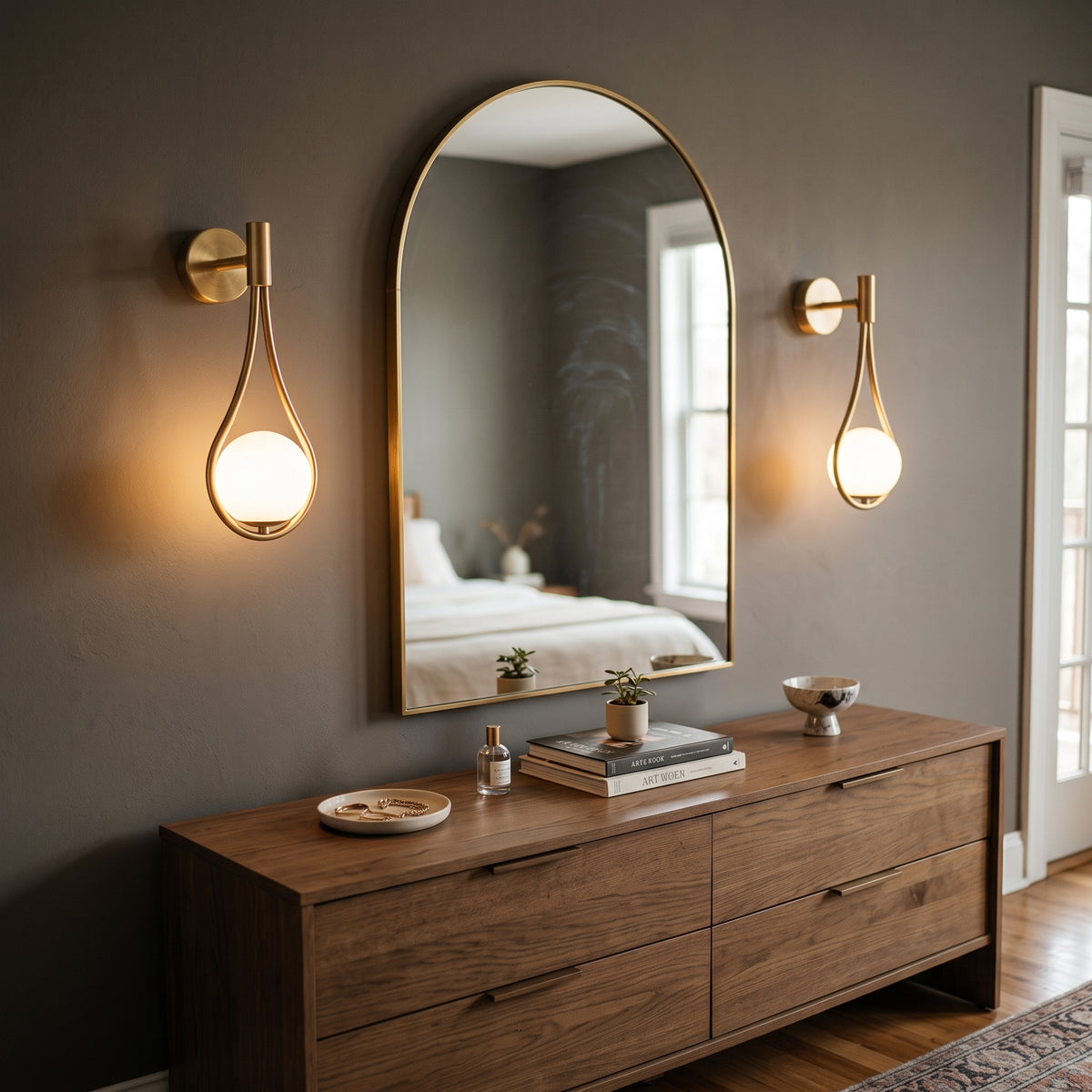 A modern bedroom scene with a large arched mirror centered on a dark gray wall, flanked by two matching gold wall sconces. Below the mirror is a wooden dresser with books, small plants, and decorative objects.