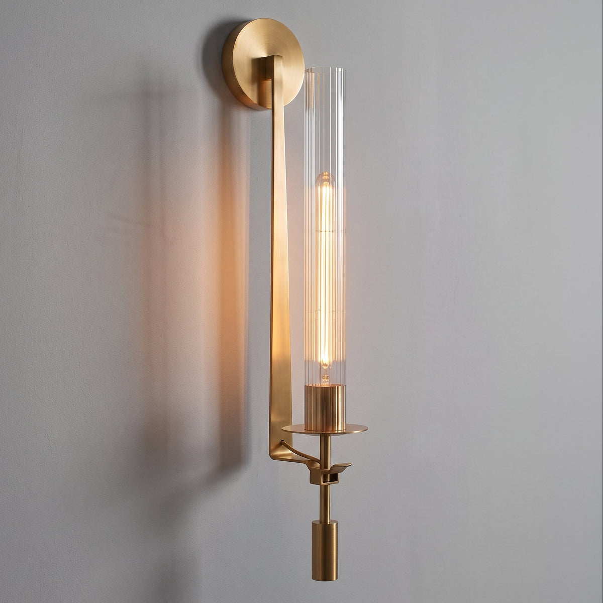A modern wall-mounted light fixture with a brushed gold metal frame and a clear cylindrical glass shade, shown illuminated against a plain light gray wall.