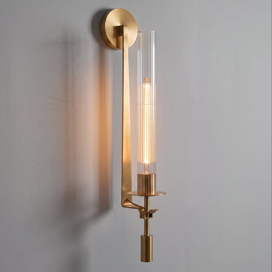 A modern wall-mounted light fixture with a brushed gold metal frame and a clear cylindrical glass shade, shown illuminated against a plain light gray wall.