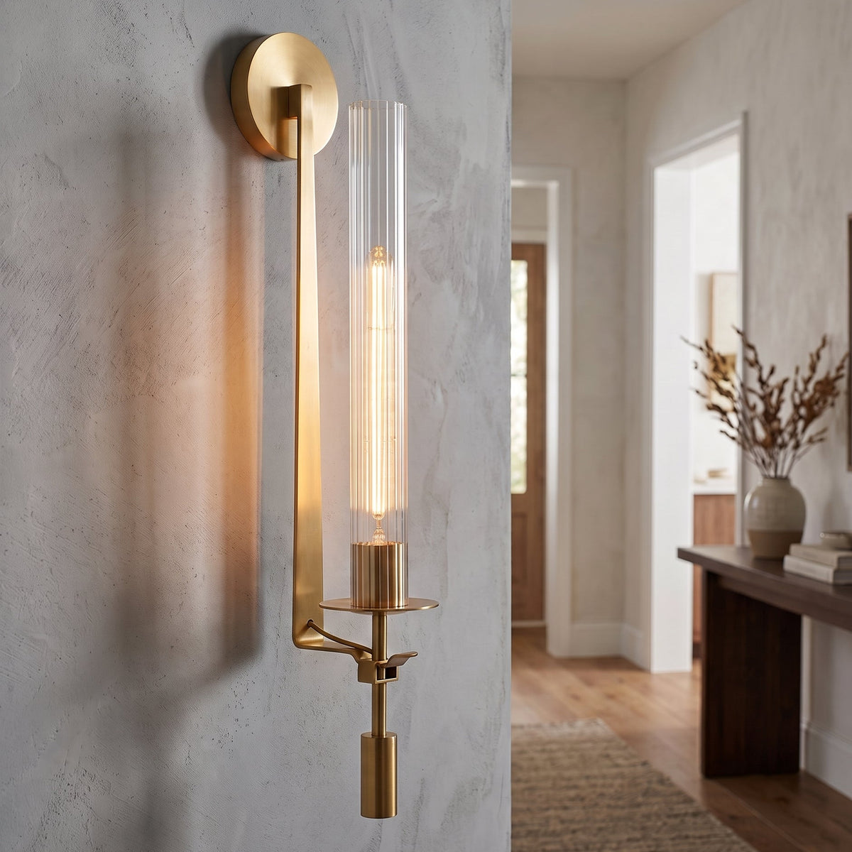 A modern wall sconce with a brushed gold circular wall mount, a vertical gold arm, and a clear cylindrical glass shade containing a warm white filament light, shown mounted on a textured light gray wall in a hallway.