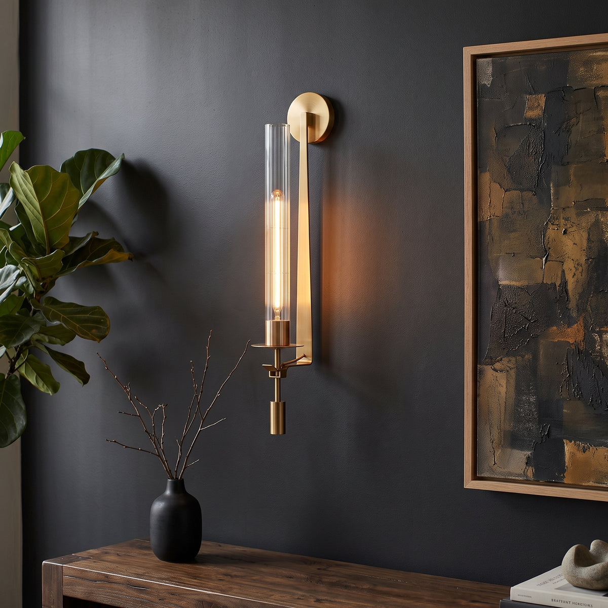A modern wall sconce with a vertical clear glass cylinder and a warm brass-toned metal body, mounted on a dark charcoal wall in a styled interior next to a plant and framed artwork.
