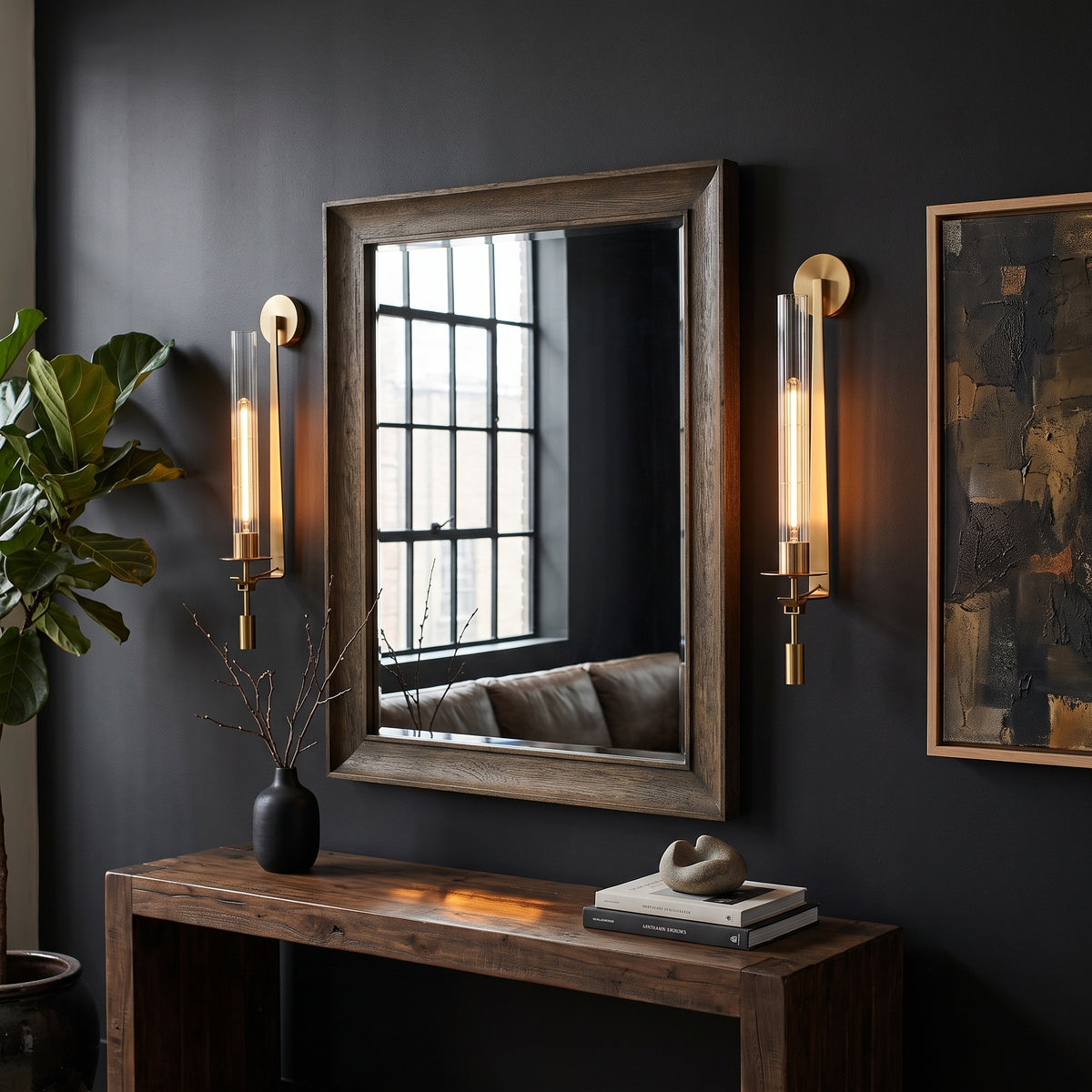 A rectangular wall mirror with a rustic dark wood frame hangs on a dark charcoal wall above a wooden console table. Two matching gold and clear glass sconces flank the mirror, and a framed abstract print is partially visible on the right.