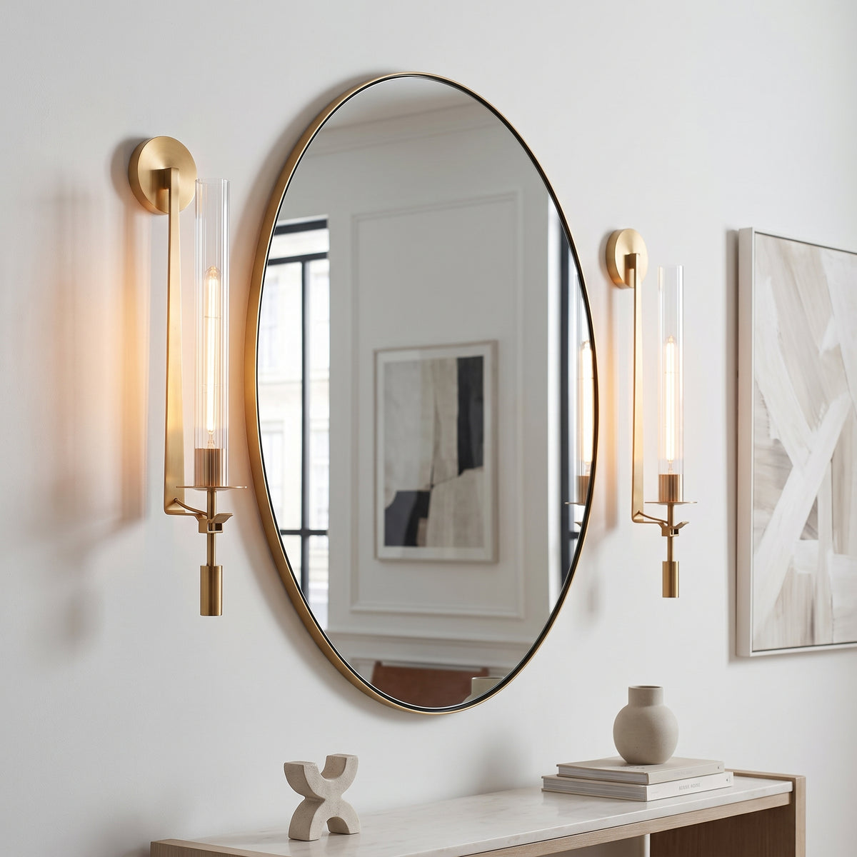 An oval wall mirror with a gold frame is mounted on a white wall, flanked by two matching gold wall sconces with clear glass tubes. A light wood console table with books and a small vase sits below.