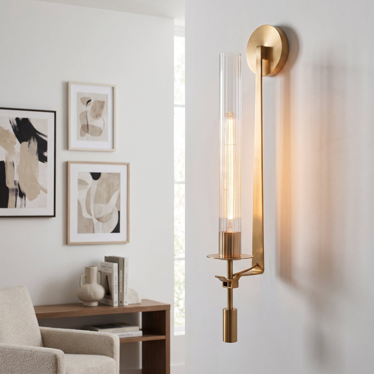 A modern wall sconce light fixture with a brushed gold metal frame and a clear cylindrical glass shade, mounted on a white wall in a bright living room setting.