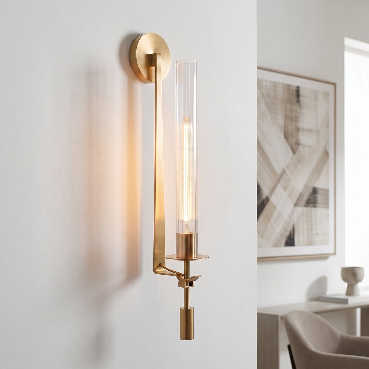A modern wall sconce with a brushed gold finish and a clear glass cylinder shade, mounted on a white wall in a contemporary interior setting.