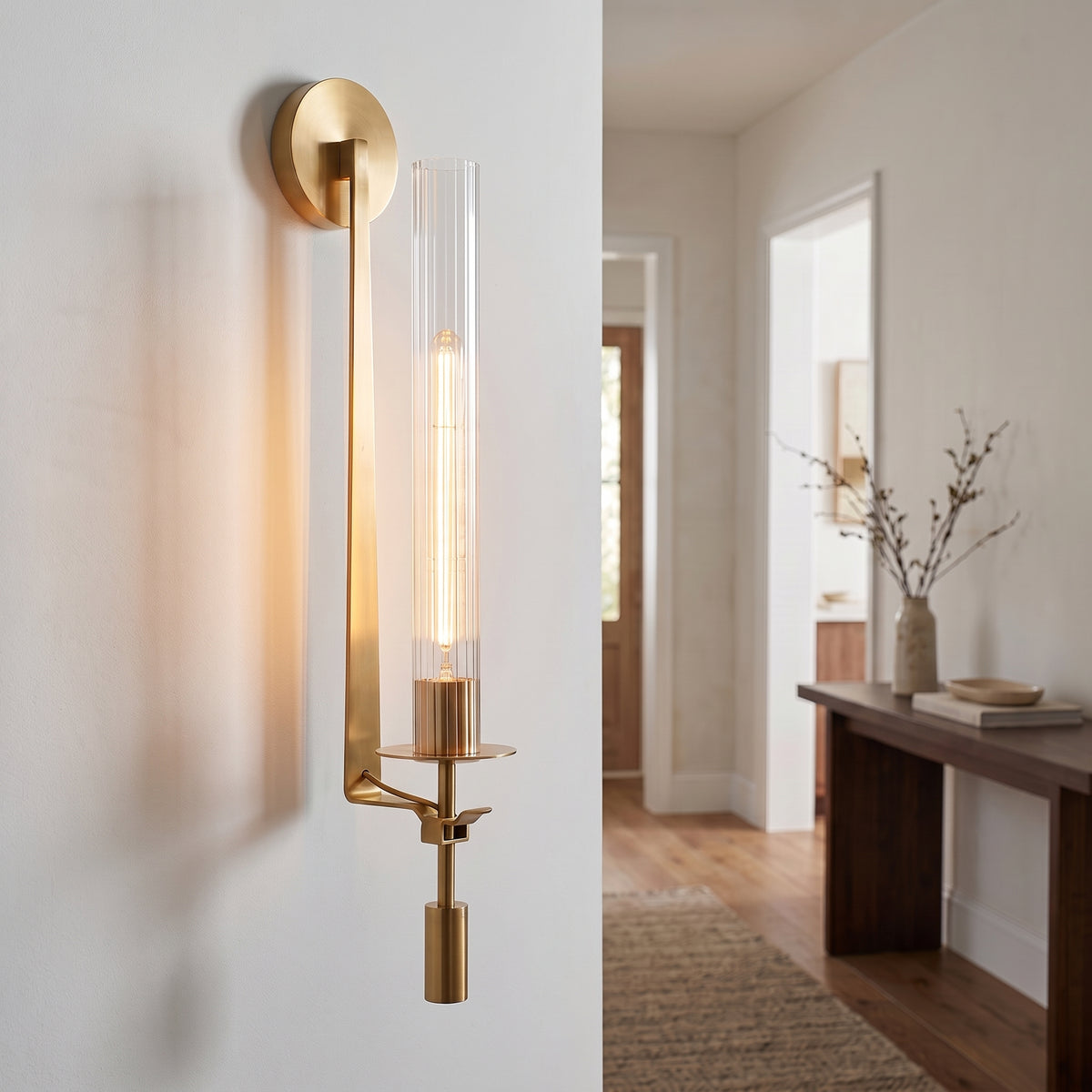 A modern wall sconce with a brushed gold finish and a clear cylindrical glass shade, mounted on a white wall in a bright interior hallway.