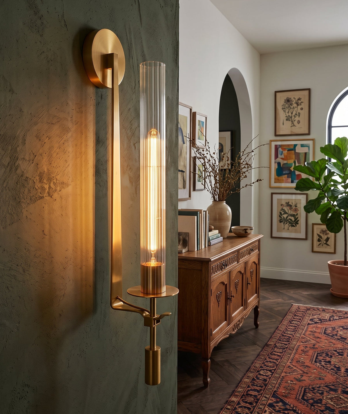 A modern wall sconce with a vertical clear glass cylinder and a warm brass/gold metal body, mounted on a textured dark green wall in a styled interior hallway with a wooden console, framed art, and a patterned rug.