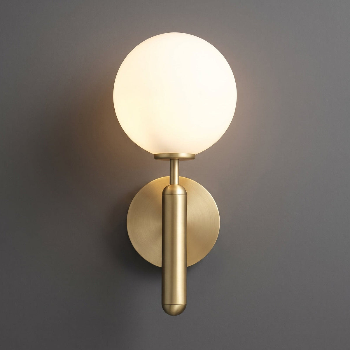 A modern wall sconce with a glowing frosted white spherical globe and a brushed brass-finished circular wall mount and vertical stem, shown against a dark gray wall.